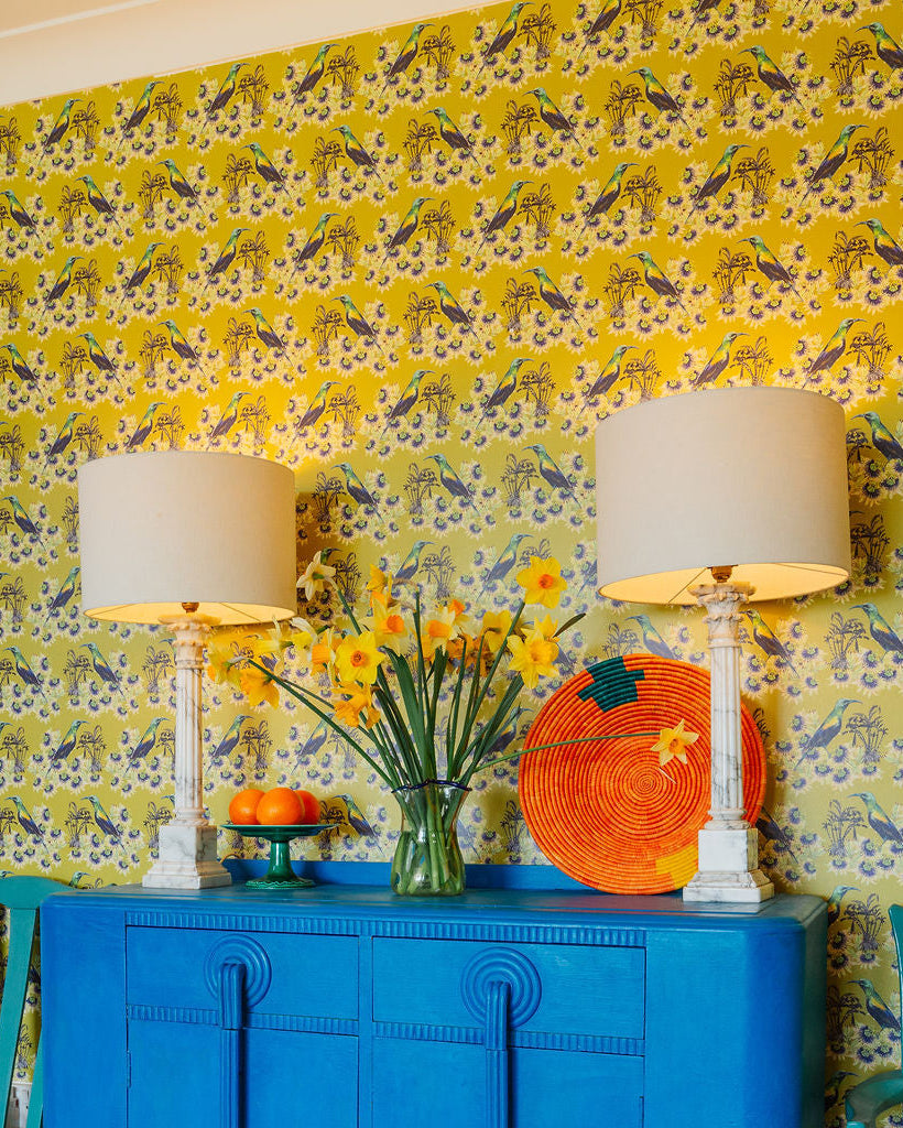 Kirimiri patterned wallpaper in Olive with a blue cabinet, orange lampshade, and spring floral arrangement.