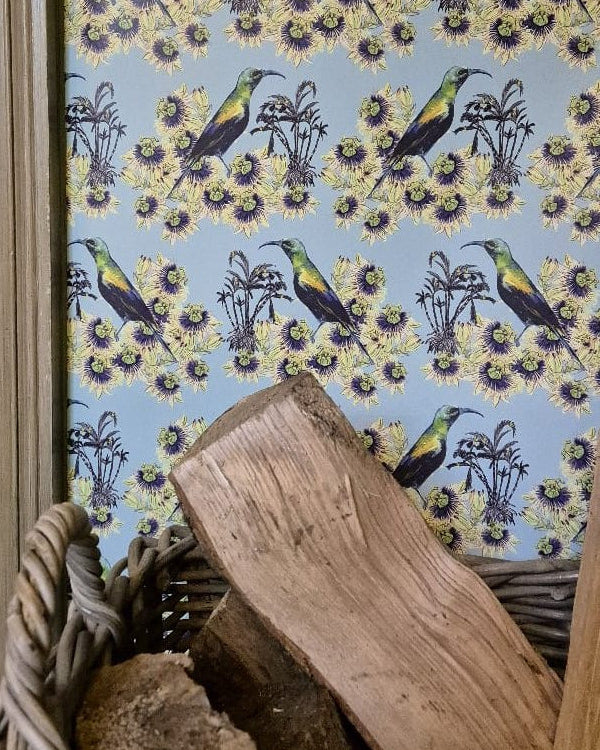 Kirimiri Wallpaper in Winter Sky (pale blue) with bird and flower pattern in a room with wooden logs and a basket.