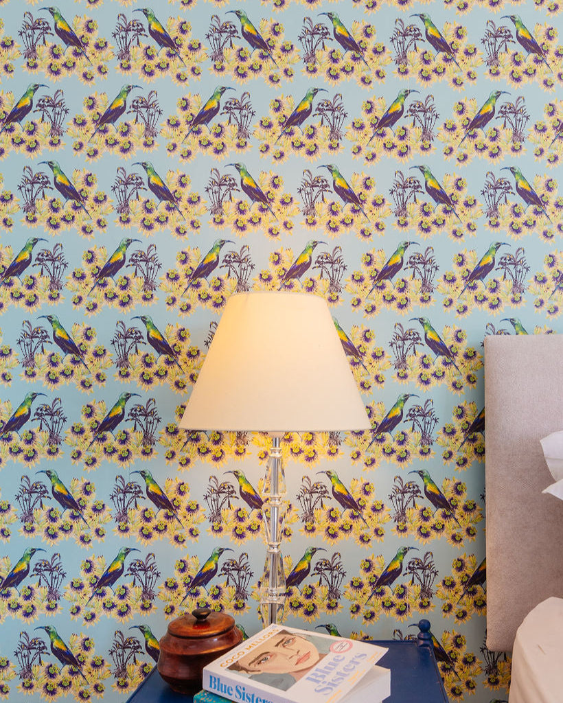 Bedroom interior with blue side table, lamp, and books against a sky blue Kirirmiri colourful tropical bird wallpaper.