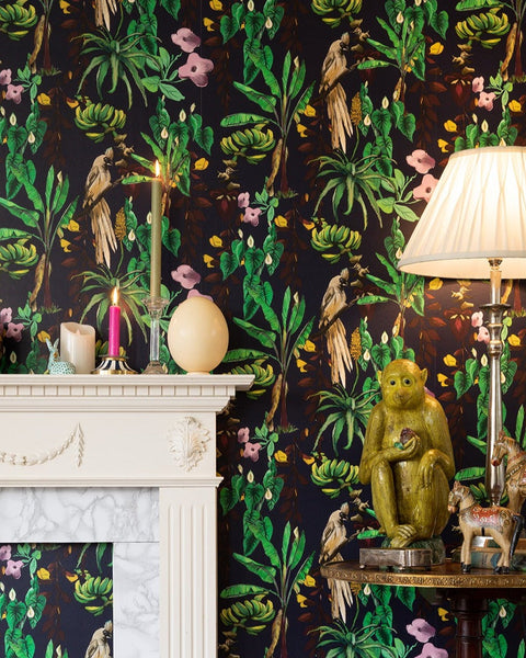 Kitito wallpaper featuring tropical plants, mousebirds on a dark blue background with a decorative mantle piece with candles, ostrich egg and decorative side table with lamp, silver box, ceramic monkey and two wooden painted folk style horses andtwo old bound books all on the side table.
