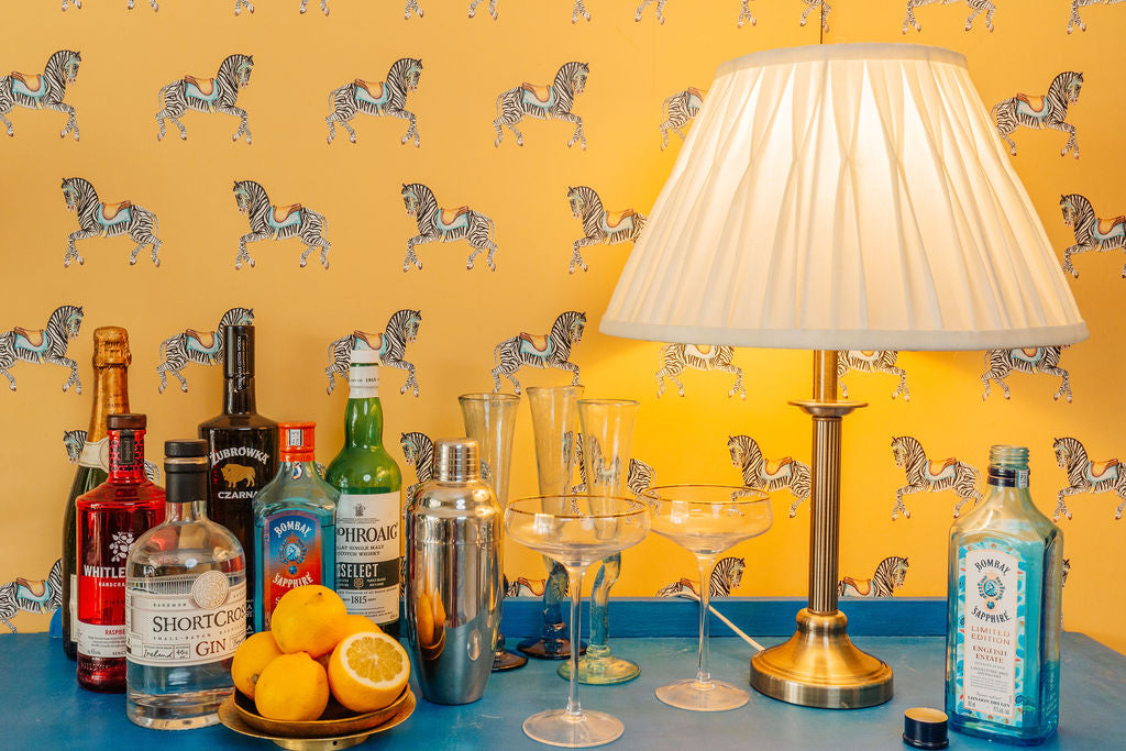 Zebra Parade Wallpaper in Moroccan Ochre (Yellow) background with a cocktail drinks and lamp on a blue surface.
