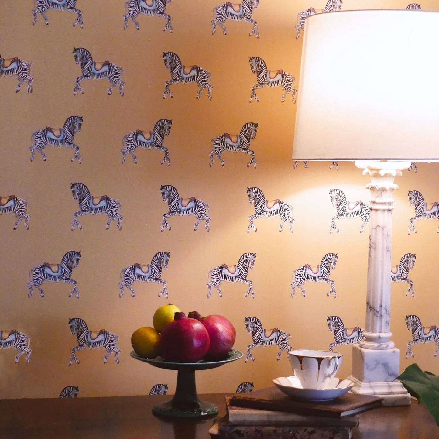 Zebra Parade Wallpaper wall with table and lamp with teacup , book sand a cake stand with fruit.
