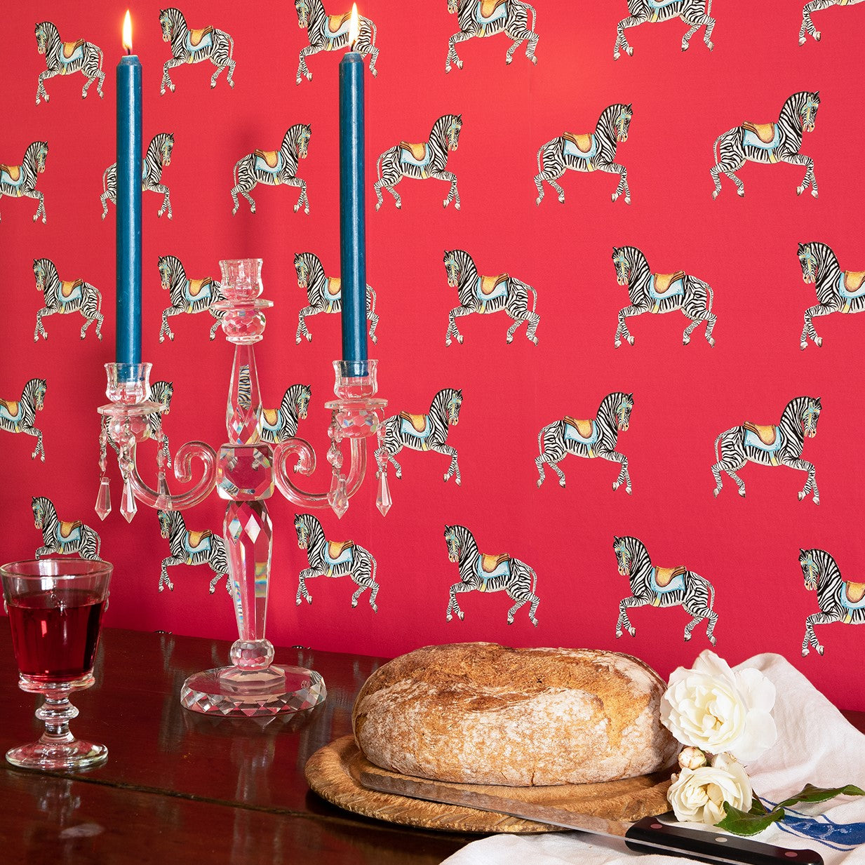 A wall adorned with raspberry red Zebra Parade wallpaper featuring a repeated zebra pattern, with a lit candle and a loaf of bread on the surface.